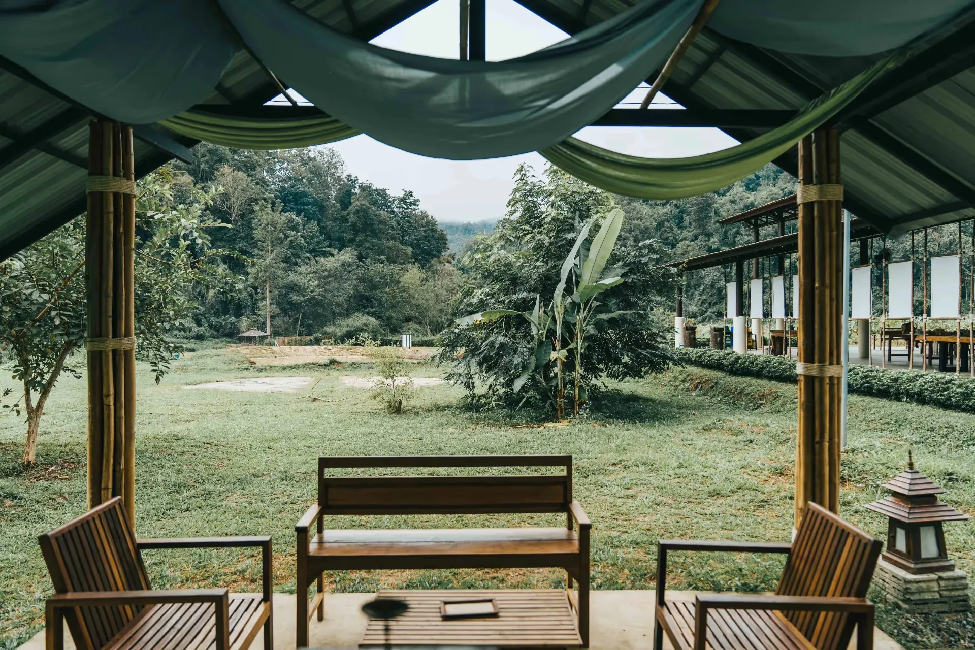 A terrace with 3 empty wooden chairs and a table in the middle. The surroundings are a garden with green trees and a long patio with a roof